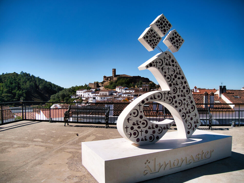 White modernist sculptural installation with carved symbols and mathematical notation, overlooking a hilltop town with castle, clear blue sky