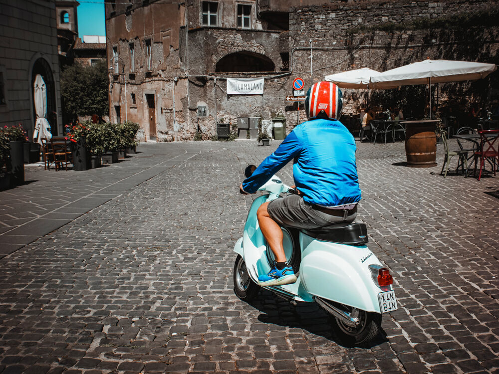 A rider in a blue jacket and striped helmet crossing a cobbled square on a pale blue Vespa, Anagni