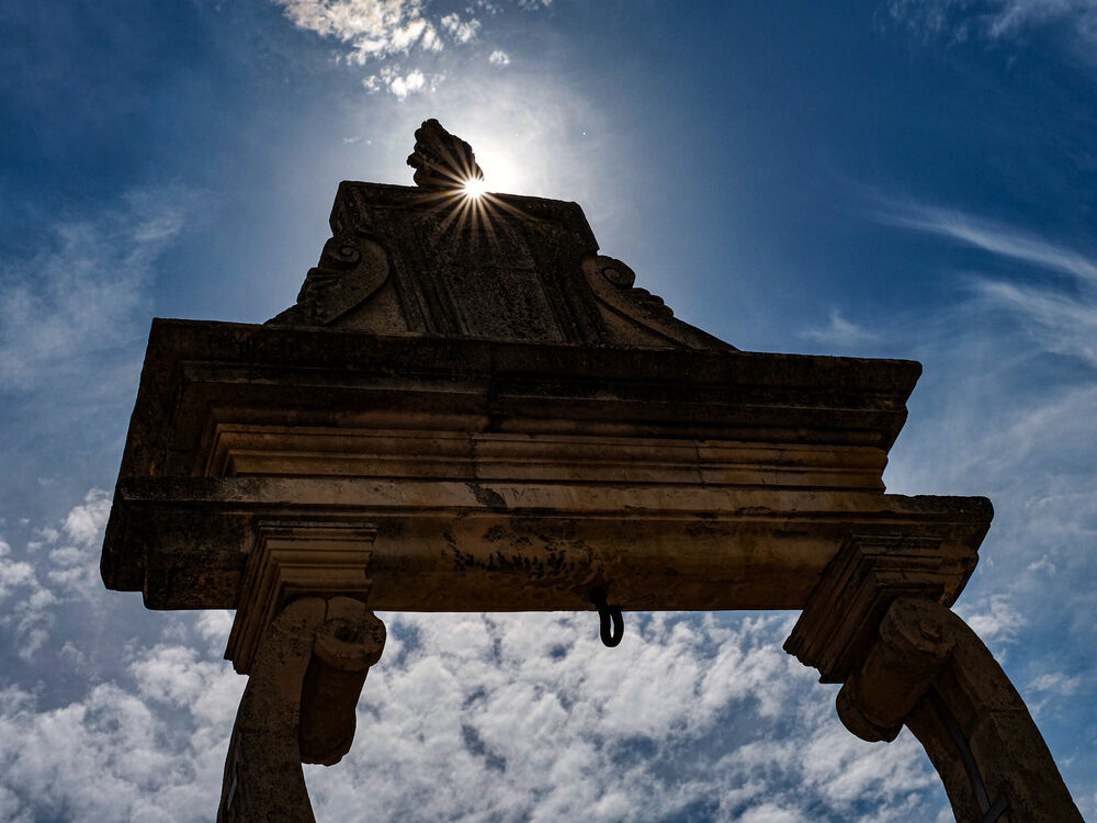A stone portal silhouetted from below with a sunstar breaking over its carved pediment, Atri