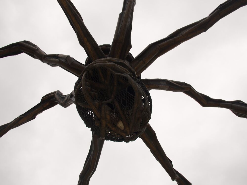 Looking straight up at the underside of the giant Maman spider sculpture, its legs radiating against an overcast sky, Bilbao
