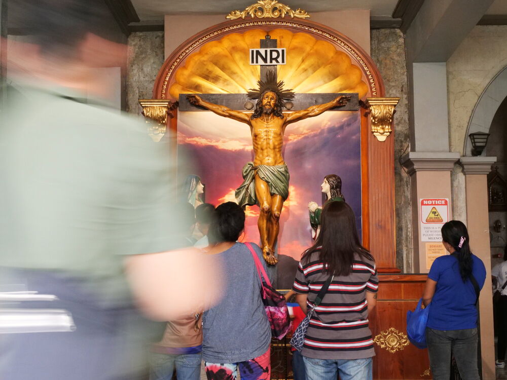 Worshippers standing before a crucifix altar while a passer-by blurs through the frame, Basilica del Santo Niño, Cebu