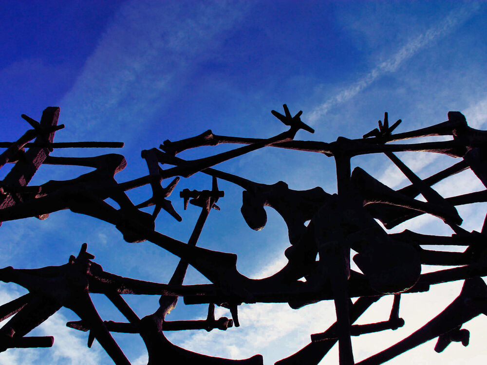 Silhouette from below of Nandor Glid's bronze memorial of emaciated figures entangled in barbed wire against a blue sky, Dachau