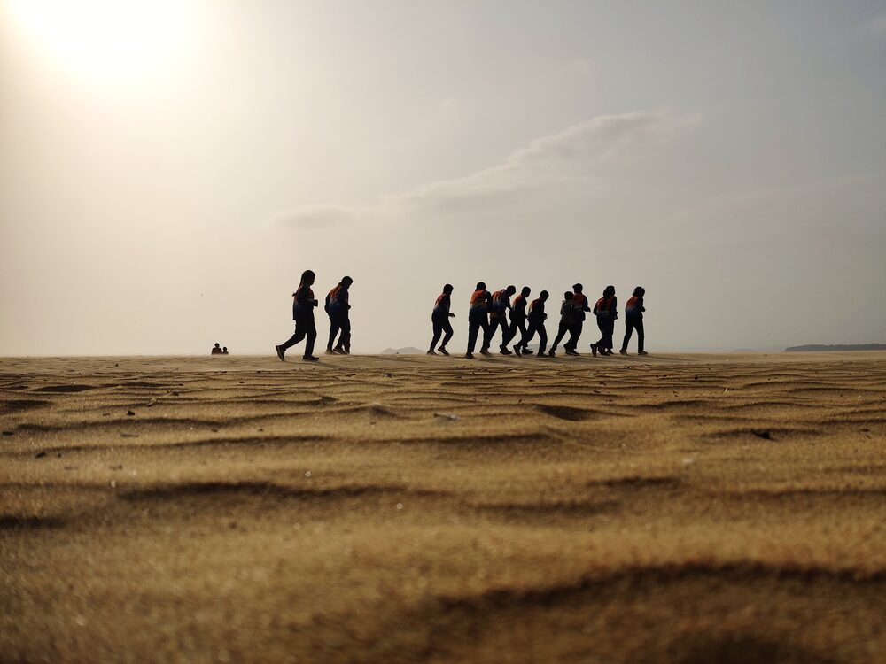 Silhouetted line of schoolchildren walking across a vast hazy beach at low light, India