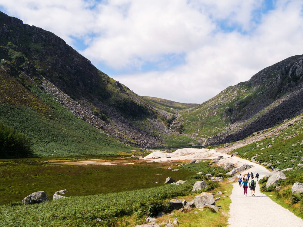Hikers walking a gravel path through the Glenealo valley at Glendalough with a distant waterfall, Ireland