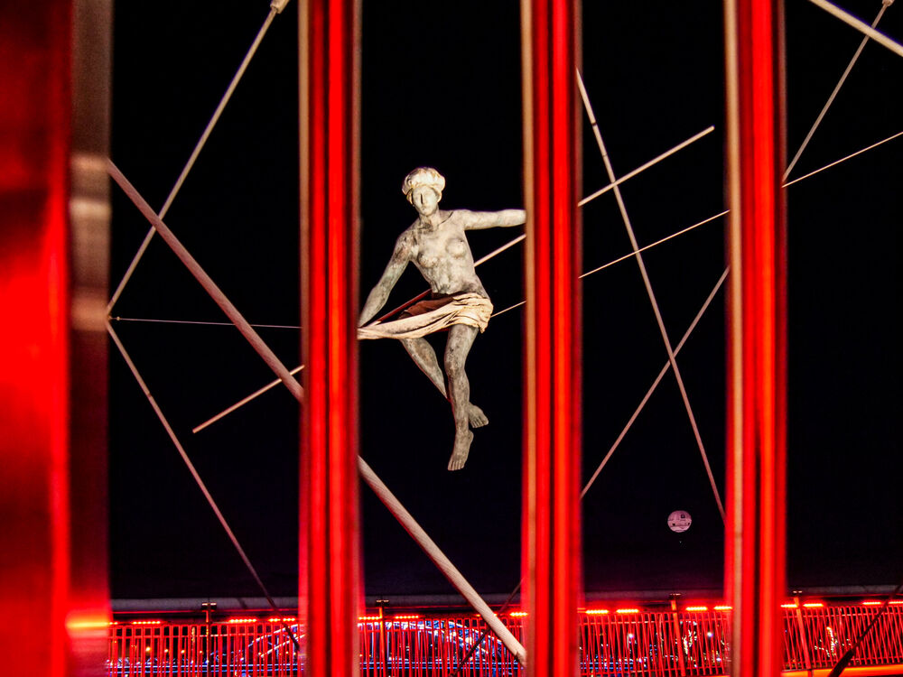 Balancing acrobat sculpture suspended among red-lit steel struts of the Bernatek footbridge at night, Kraków