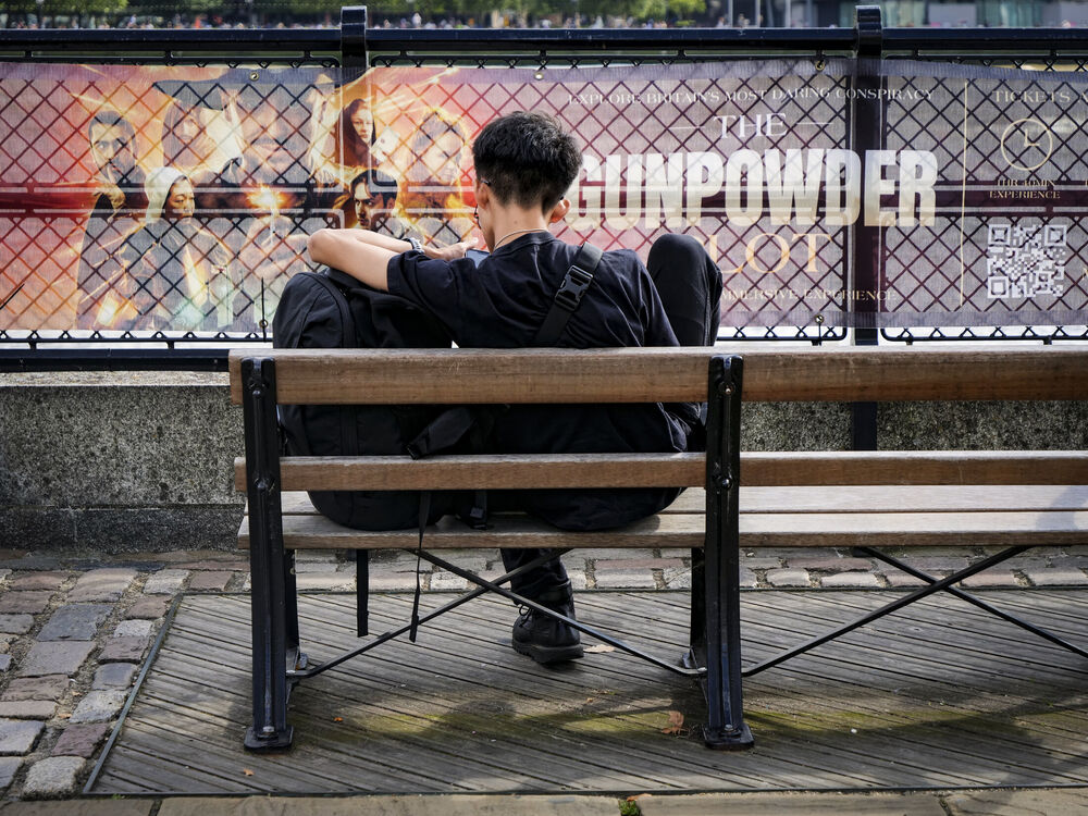 A young man seen from behind sitting on a bench checking his phone in front of a Gunpowder Plot banner, London