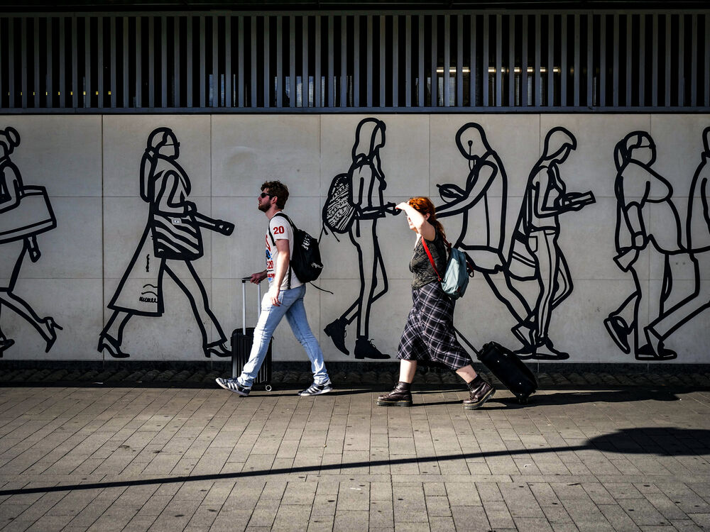 A man and a woman pulling suitcases past a wall frieze of black line-art walking figures, London