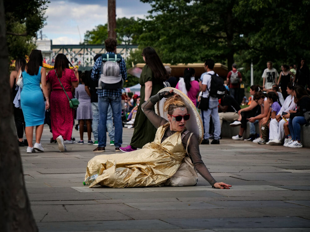 A street performer in a gold dress and red sunglasses reclining mid-act on the South Bank pavement, London