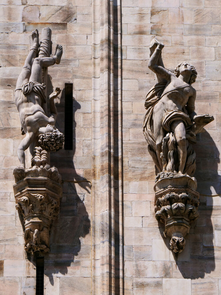 Two marble statues on carved brackets against the wall of Milan Cathedral, one inverted, with hard shadows