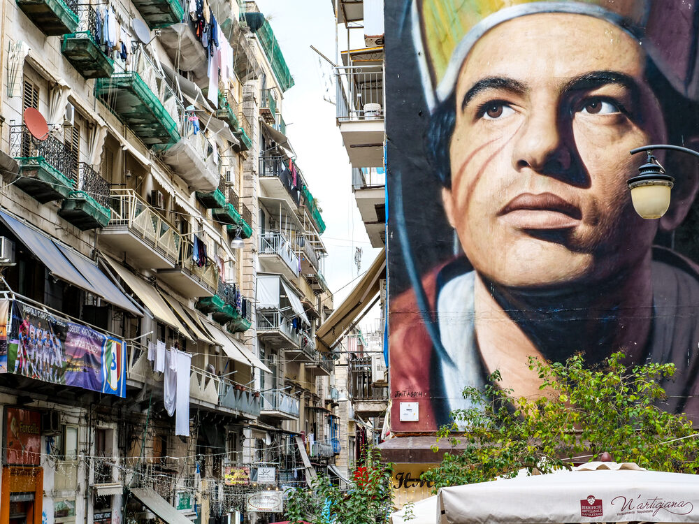 Large street mural of San Gennaro towering over a narrow Naples street hung with laundry