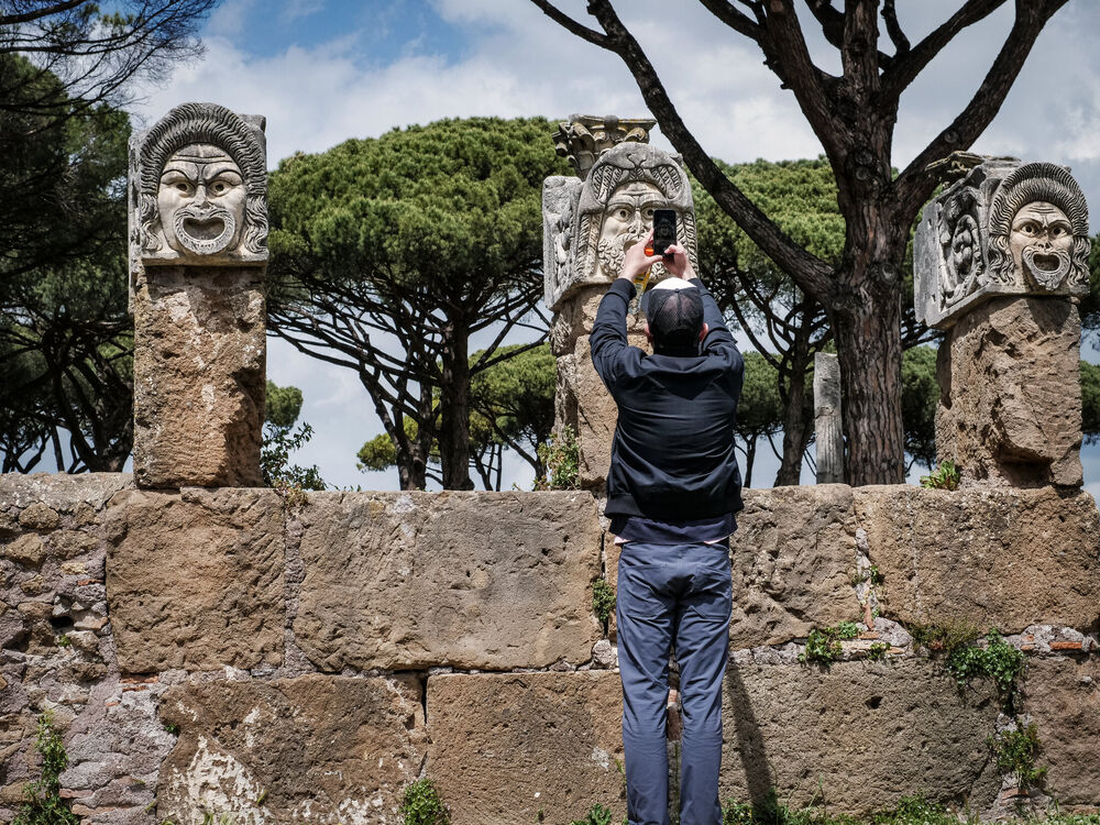 A man photographing giant carved marble theatre masks on pillars among umbrella pines at Ostia Antica