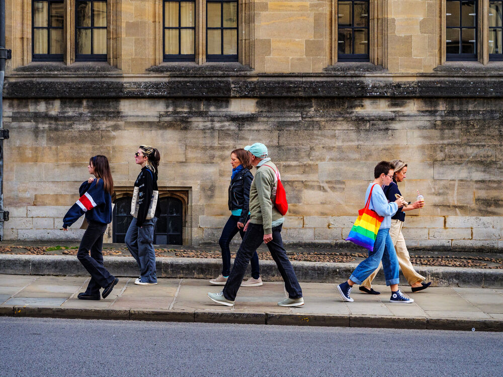 Six pedestrians walking in profile past a weathered stone college wall in Oxford, one carrying a rainbow tote bag