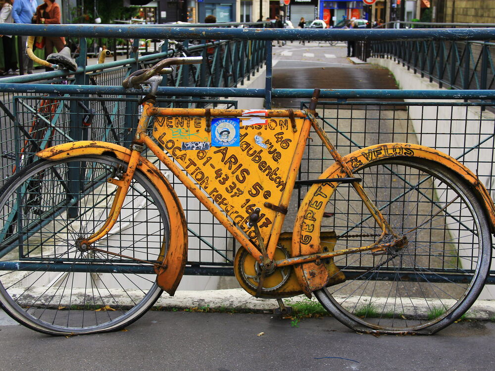 An old yellow bicycle painted with a bike shop's hand-lettered advertisement, chained to a green railing in Paris