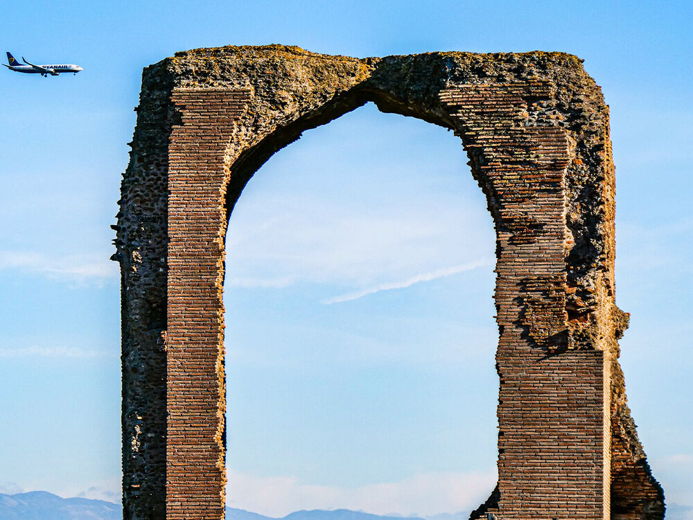 A Ryanair jet on approach passing behind a ruined Roman brick arch against blue sky, Rome