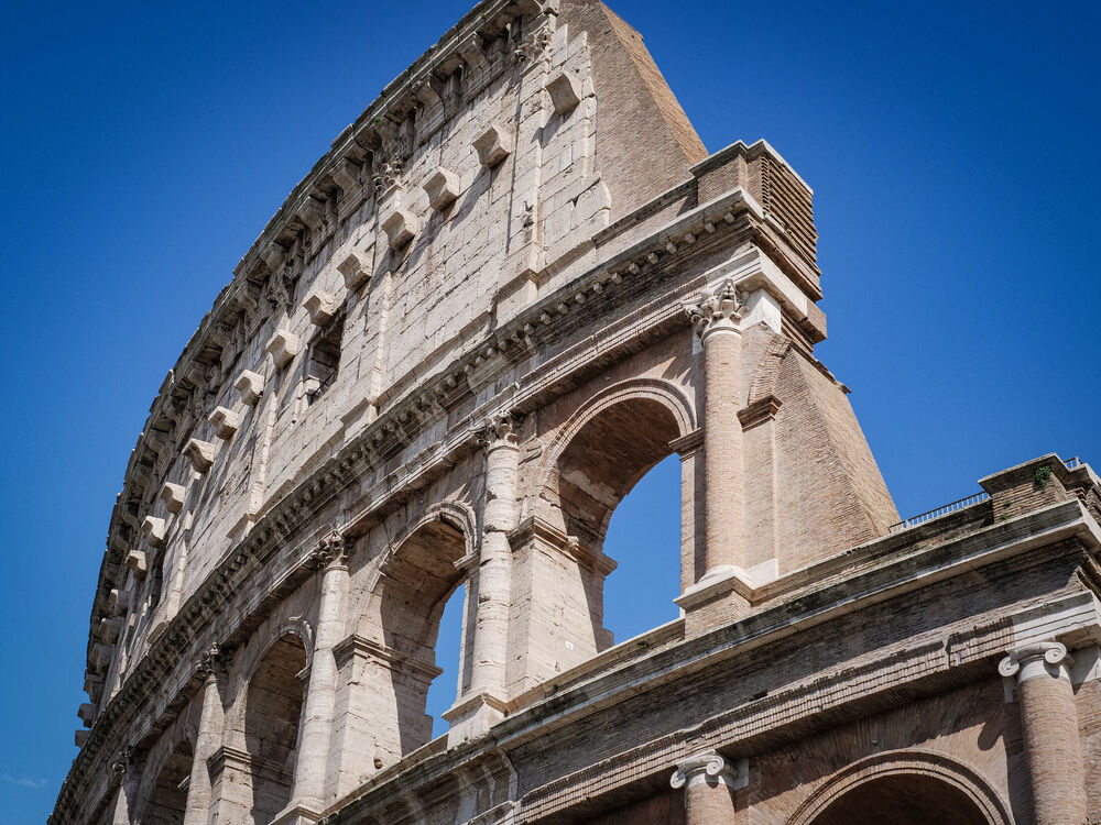 Looking up at the curved upper arches and corbels of the Colosseum against deep blue sky, Rome