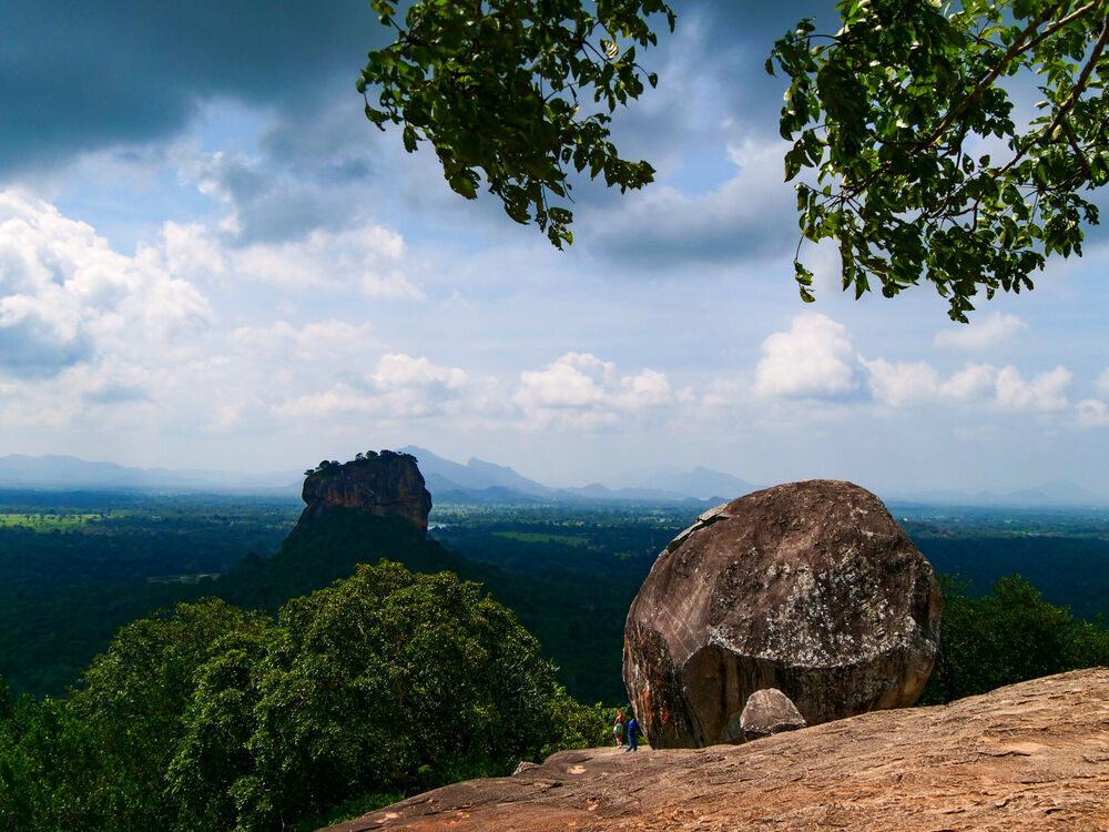 Sigiriya rock fortress rising from forested plains seen from Pidurangala rock with a boulder in the foreground, Sri Lanka