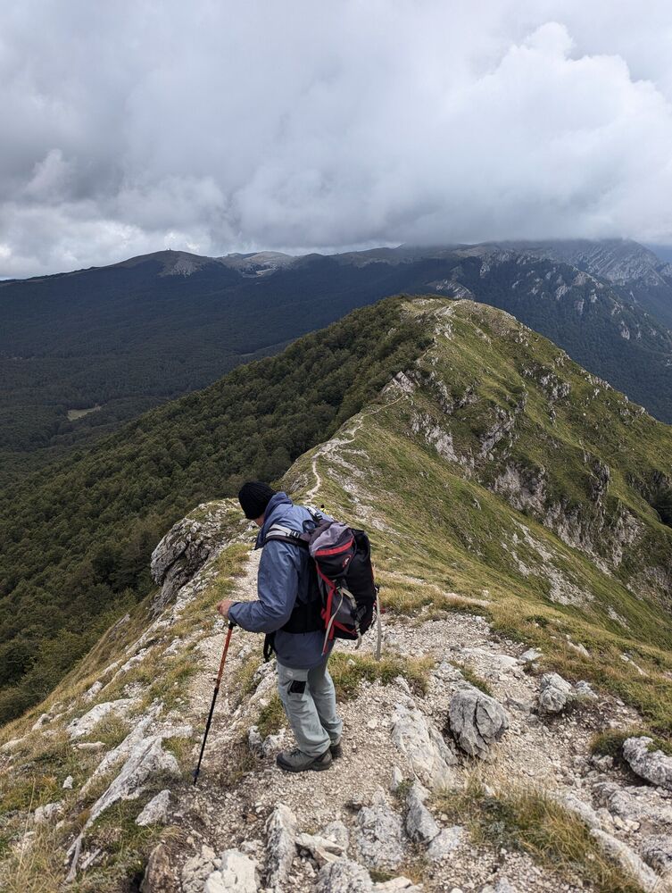 A hiker with a backpack descending a narrow mountain ridge path in the Apennines under low cloud
