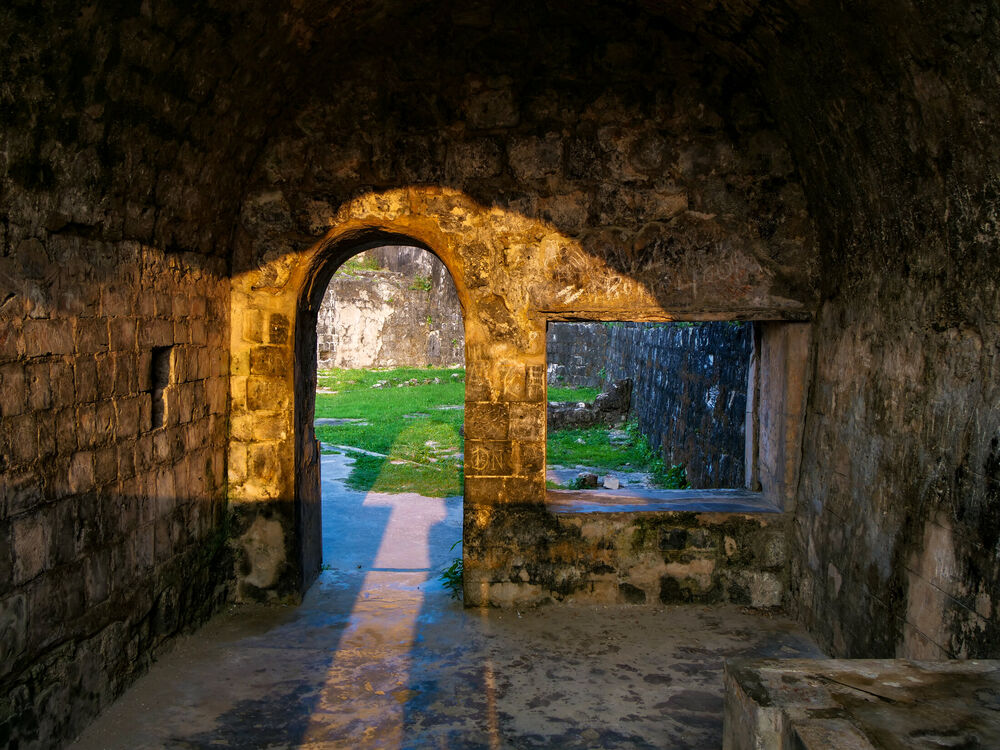 Golden evening light striking an arched doorway inside a dark stone fort casemate with a grass courtyard beyond, Sri Lanka