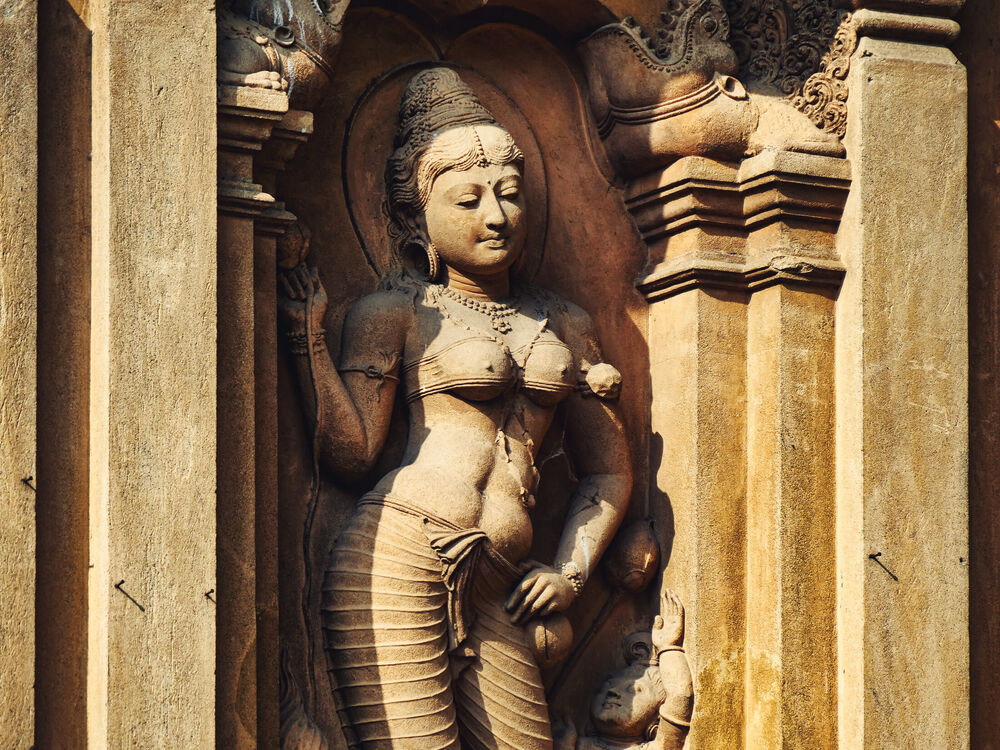 A carved stone relief of a serene female figure in a niche on a Sri Lankan temple wall, warm afternoon light