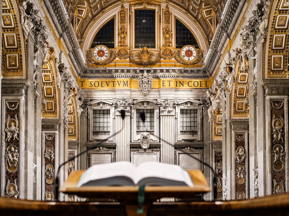 An open lectionary on a lectern facing the gilded apse and twin clocks inside St Peter's Basilica