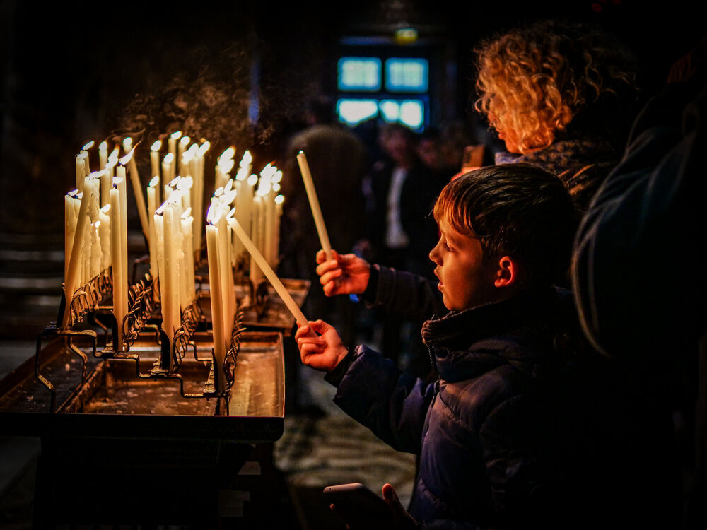 A young boy lighting a taper candle at a votive candle rack inside a dark Venetian church