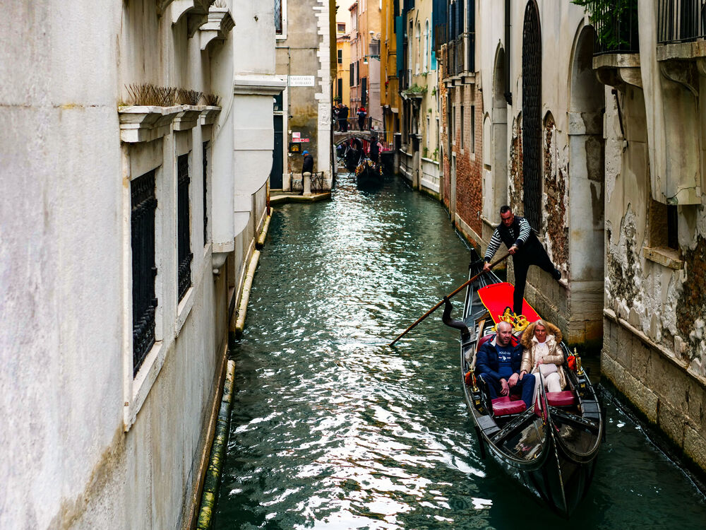 A gondolier pushing off a canal wall with his foot while steering passengers down a narrow Venetian canal