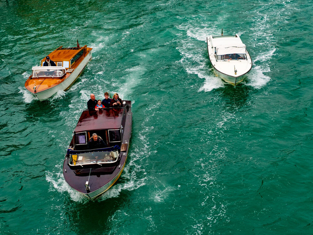 Three wooden water taxis crossing paths on green canal water seen from above, Venice